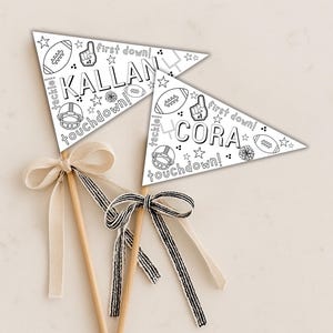 Puede incluir: Dos banderines con temática de fútbol sobre palos de madera, cada uno con un lazo. Un banderín dice "KALLAN" y el otro "CORA", con gráficos de fútbol y las palabras "first down" y "touchdown". Los lazos son crema y a rayas blancas y negras.