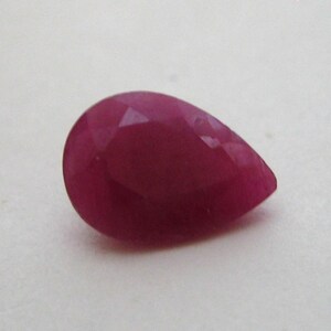 May include: A pear-shaped, faceted gemstone with a deep magenta-red color. The stone features a smooth, polished surface with multiple triangular facets that reflect light, creating a clear, translucent appearance with a pointed end.