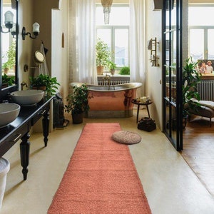 May include: A salmon-colored bath mat with a textured surface is placed in front of a copper bathtub in a modern bathroom. The bathroom has a large window with white curtains and a mirror on the wall.