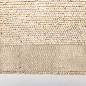 May include: Close-up of a beige rug with a textured, looped pile. The lower section features a woven, flat-weave design. The rug's edge is visible, showing a finished border. The rug is a neutral color, suitable for various interior styles.