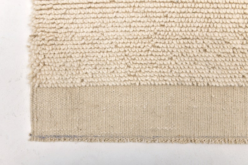 May include: Close-up of a beige rug with a textured, looped pile. The lower section features a woven, flat-weave design. The rug's edge is visible, showing a finished border. The rug is a neutral color, suitable for various interior styles.