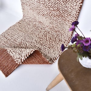 May include: A textured, brown and cream-colored rug with a geometric pattern. The rug is folded over, revealing a contrasting brown border. A small table with a vase of purple flowers is in the background.