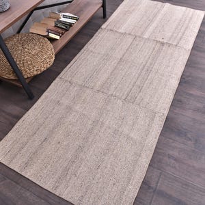 May include: A rectangular, beige rug with a textured surface. The rug is made of woven material and has a simple, neutral design. The rug is placed on a dark wood floor.