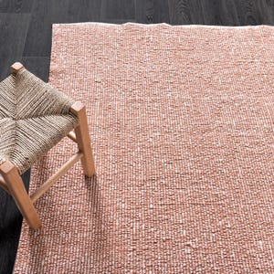 Puede incluir: Un taburete de paja trenzada con un marco de madera natural se encuentra sobre una alfombra texturizada rosa y blanca.