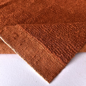 May include: Close-up of a rust-coloured woven fabric with a textured surface. The fabric is folded over, revealing the underside.