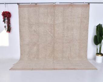 May include: A large, neutral-toned rug with a textured surface hangs from a metal rod. The rug is beige with a subtle pattern. A hanging red floral arrangement and a potted plant with green leaves are on either side.