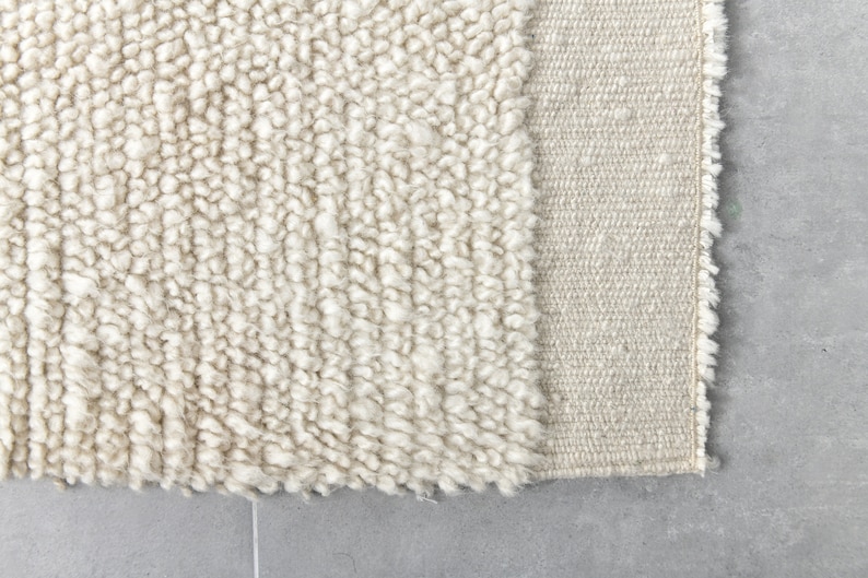 May include: Close-up of a cream-colored rug with a textured, fluffy surface. The rug has a woven edge.