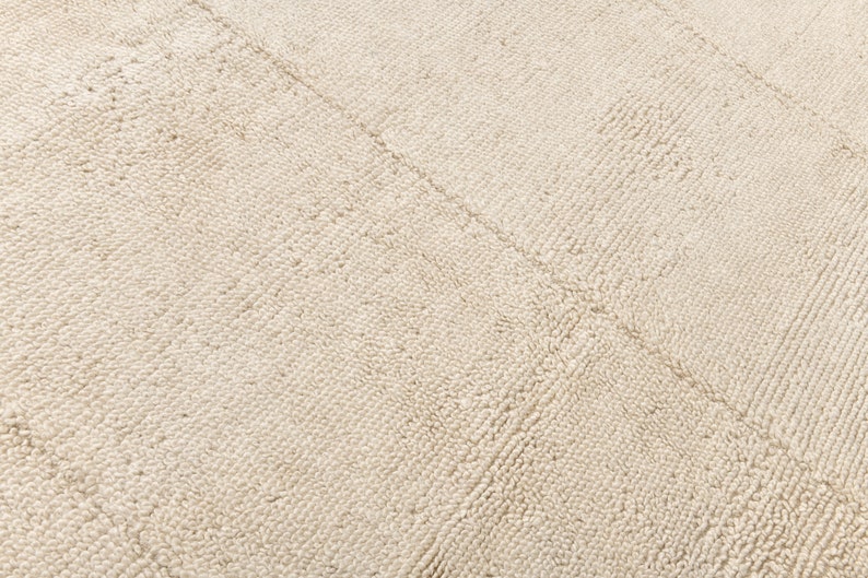 May include: Close-up of a cream-colored rug with a textured, looped pile. The rug features a subtle, geometric pattern created by the direction of the loops, with diagonal lines dividing the surface.