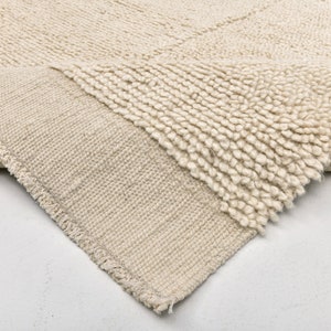 May include: Cream-colored rug with a geometric pattern. The rug has a textured, looped pile on the top surface and a woven backing. The edge of the rug is frayed, revealing the woven construction. The rug is folded over, showing the underside.