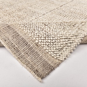 May include: Close-up of a beige and white hand-woven rug with a textured surface. The rug has a fringe border.