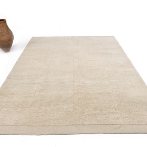 May include: A rectangular beige rug with a textured surface and a lighter border. A brown, antique-style vase is positioned to the left of the rug. The rug has a simple, neutral design.