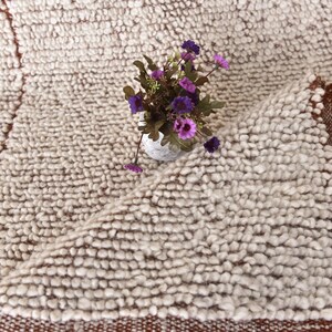 May include: A white fluffy rug with a brown border. The rug is made of small, round balls of yarn. A small bouquet of purple flowers is sitting on the rug.