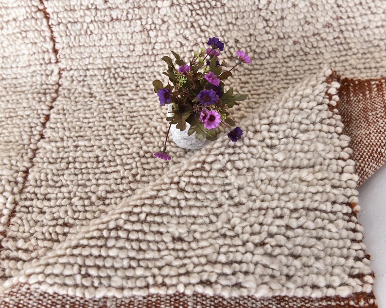 May include: A white fluffy rug with a brown border. The rug is made of small, round balls of yarn. A small bouquet of purple flowers is sitting on the rug.