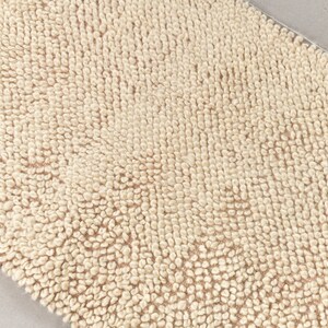 Puede incluir: Primer plano de una alfombra de ba&ntilde;o beige con una superficie texturizada. La alfombra est&aacute; hecha de un material suave y absorbente.