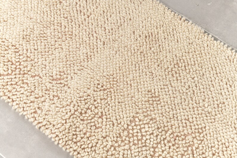 Puede incluir: Primer plano de una alfombra de ba&ntilde;o beige con una superficie texturizada. La alfombra est&aacute; hecha de un material suave y absorbente.