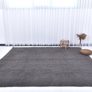 May include: A dark gray shag rug with a subtle sheen. The rug is laid out on a white floor in a room with white walls and sheer white curtains.