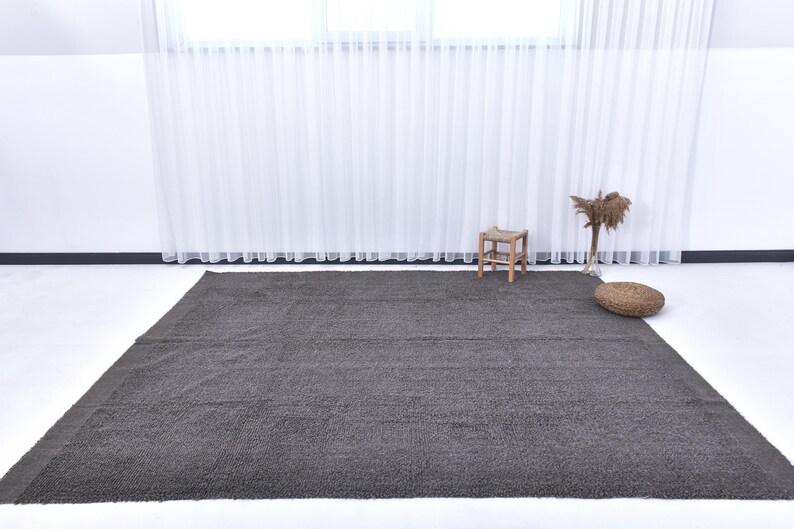 May include: A dark gray shag rug with a subtle sheen. The rug is laid out on a white floor in a room with white walls and sheer white curtains.