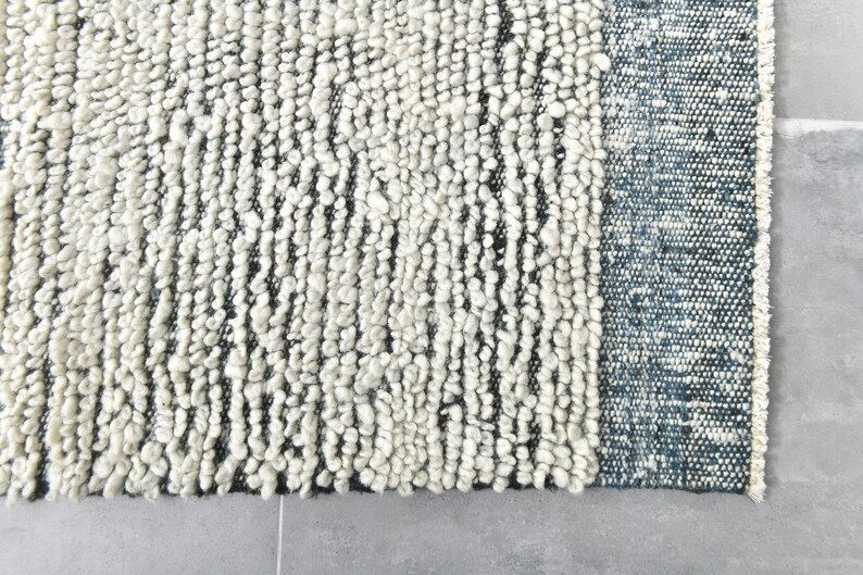 May include: Close-up of a white and gray textured rug with a blue border. The rug has a unique, hand-woven look.