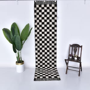 May include: Black and white checkered rug with fringe. The rug is hanging on a white wall.