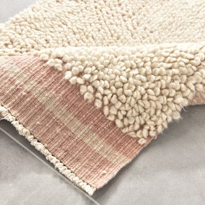 Puede incluir: Una alfombra redonda con una textura esponjosa de color crema y un borde tejido rosa. La alfombra est&aacute; doblada por la mitad, mostrando la textura y el borde.