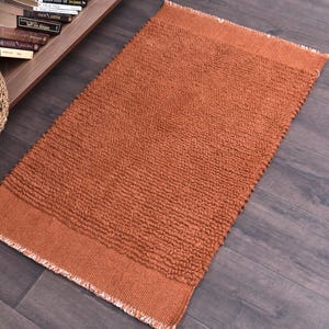 May include: A rectangular, rust-colored bath mat with a textured, looped pile. The mat has a solid border and fringed edges. The rug is placed on a dark wood floor, with a woven basket and a bookshelf visible in the background.