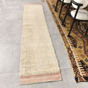 Puede incluir: Una alfombra larga, de color blanco roto y esponjosa, con un borde rosa y blanco. La alfombra est&aacute; colocada sobre un suelo de baldosas grises junto a una alfombra estampada.