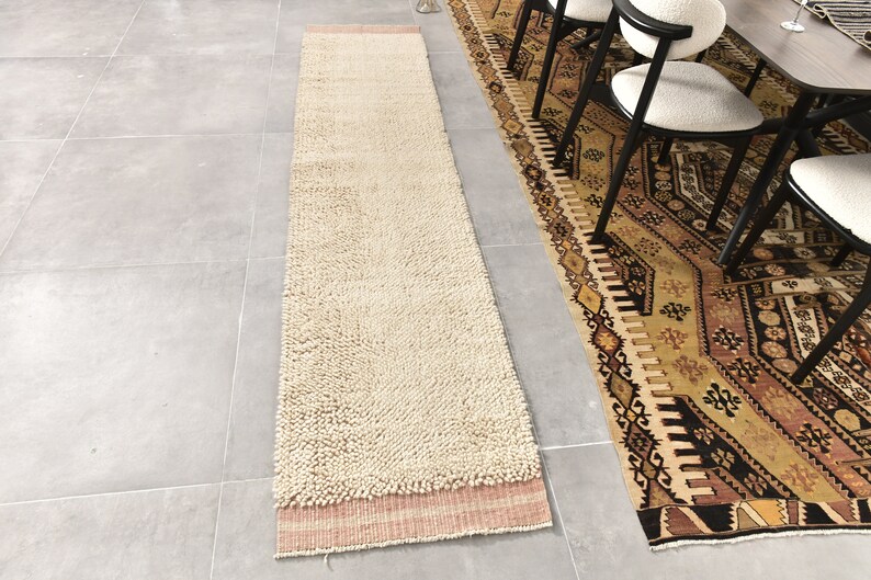 Puede incluir: Una alfombra larga, de color blanco roto y esponjosa, con un borde rosa y blanco. La alfombra est&aacute; colocada sobre un suelo de baldosas grises junto a una alfombra estampada.