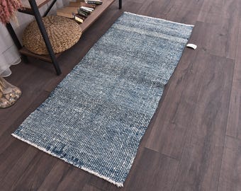 Può includere: Un tappeto rettangolare blu e bianco con una trama strutturata. Il tappeto è posato su un pavimento in legno scuro. Il design del tappeto presenta un aspetto sbiadito e invecchiato, che si aggiunge alla sua estetica vintage. Il tappeto è uno stile runner.