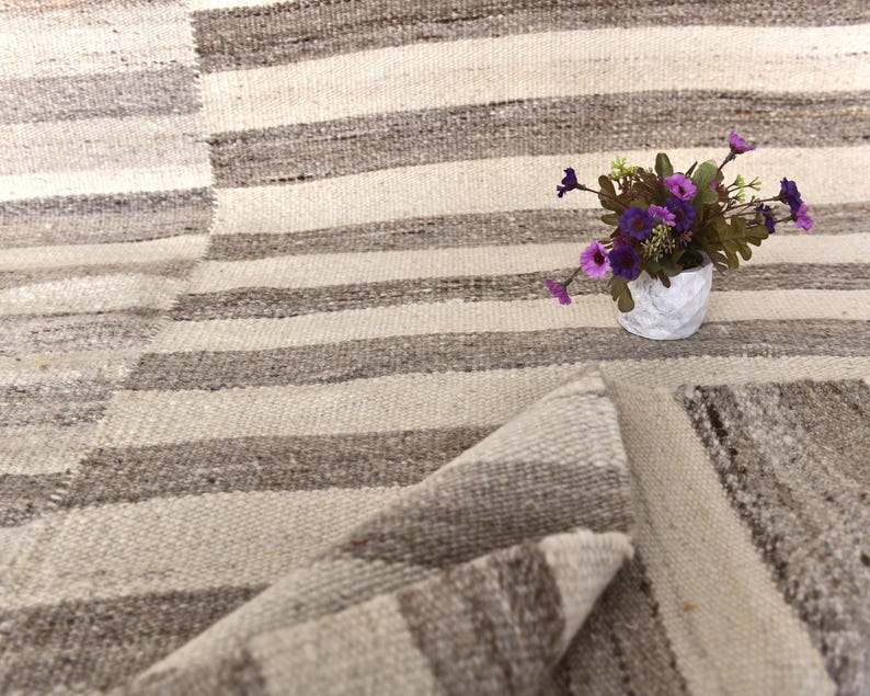 Pu&ograve; includere: Un tappeto a strisce bianche e marroni con un bordo piegato. Un piccolo vaso bianco con fiori viola &egrave; sul tappeto.