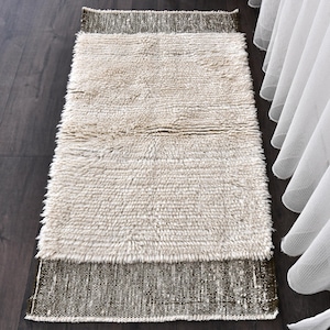 Puede incluir: Una alfombra rectangular beige con una textura peluda y desgreñada. La alfombra tiene un borde marrón más oscuro en cada extremo. La alfombra está colocada sobre un suelo de madera oscura, con cortinas blancas visibles al fondo.