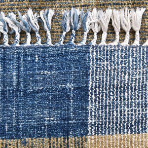 May include: Close-up of a blue and tan woven rug with a textured design. The rug features a blue section with a geometric pattern, a tan border, and white fringe along the top edge. The rug's texture is visible.