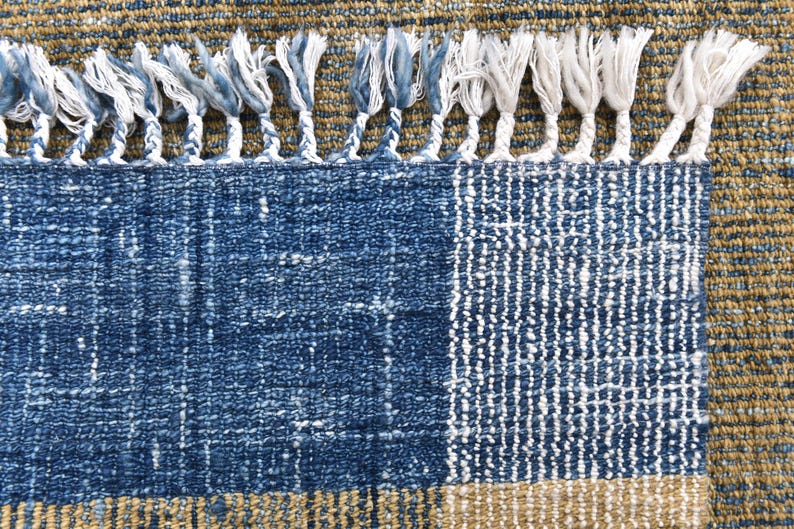 May include: Close-up of a blue and tan woven rug with a textured design. The rug features a blue section with a geometric pattern, a tan border, and white fringe along the top edge. The rug's texture is visible.