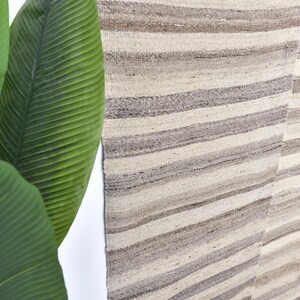 Pu&ograve; includere: Un primo piano di un tappeto tessuto con un motivo a strisce in tonalit&agrave; beige e grigio. Il tappeto ha un aspetto e una sensazione naturali e rustici.