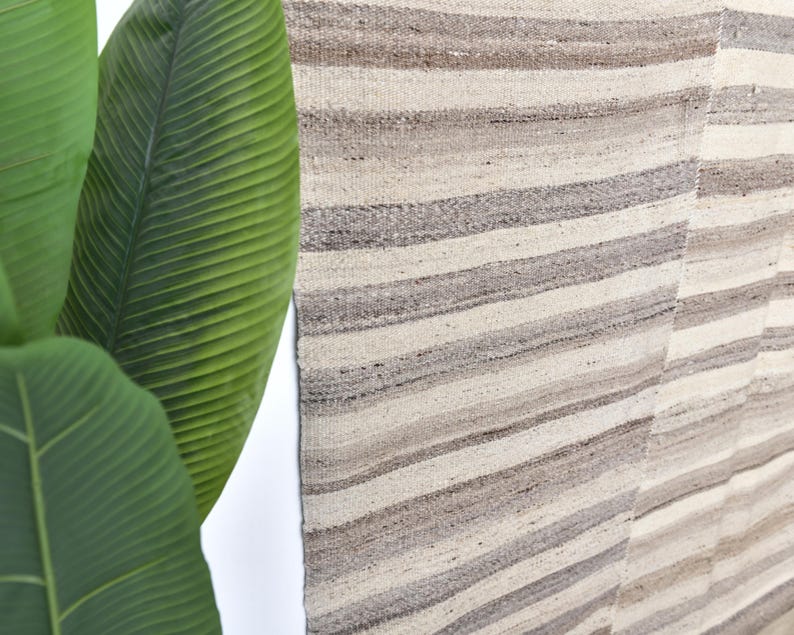 Pu&ograve; includere: Un primo piano di un tappeto tessuto con un motivo a strisce in tonalit&agrave; beige e grigio. Il tappeto ha un aspetto e una sensazione naturali e rustici.