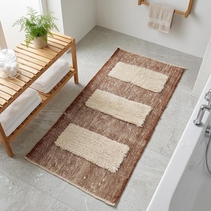 Puede incluir: Escena de baño con una alfombra rectangular marrón con tres acentos rectangulares color crema. Un banco de madera sostiene toallas blancas, una planta y artículos de tocador. También se ven una bañera blanca y un toallero.