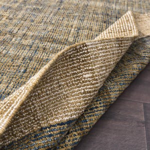 May include: Close-up of a woven rug with a textured surface. The rug features a blend of tan, blue, and white threads, creating a patterned design. The edge of the rug is folded over, revealing the underside.