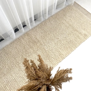 May include: A natural fiber rug in a light beige color. The rug has a textured, woven appearance and is laid out on a white surface.