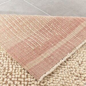 Puede incluir: Primer plano de una alfombra esponjosa de color crema con un borde rayado rosa y crema. La alfombra est&aacute; doblada por la mitad, mostrando la parte inferior de la alfombra.