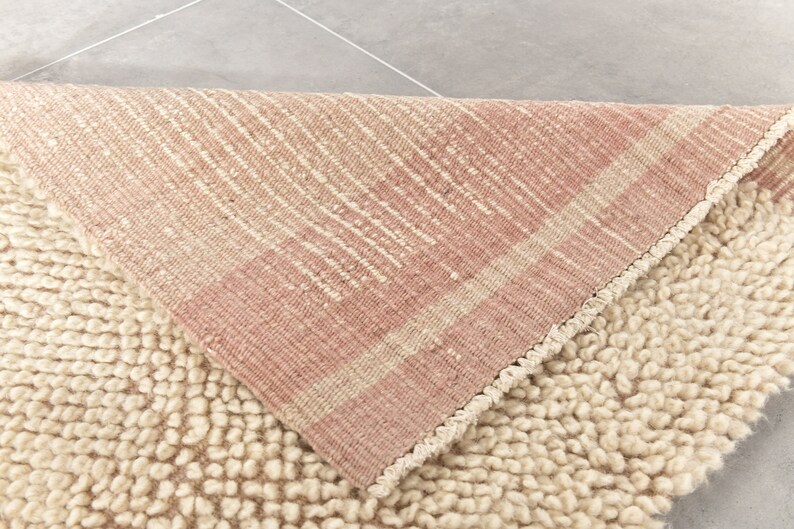 Puede incluir: Primer plano de una alfombra esponjosa de color crema con un borde rayado rosa y crema. La alfombra est&aacute; doblada por la mitad, mostrando la parte inferior de la alfombra.