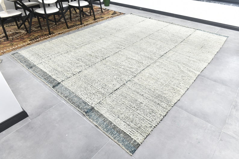 May include: A white and gray area rug with a blue and gray border. The rug has a textured, woven look and is laid out on a gray tiled floor.