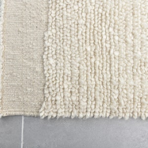 May include: Close-up of a cream-colored rug with a textured, fluffy surface. The rug has a woven edge.