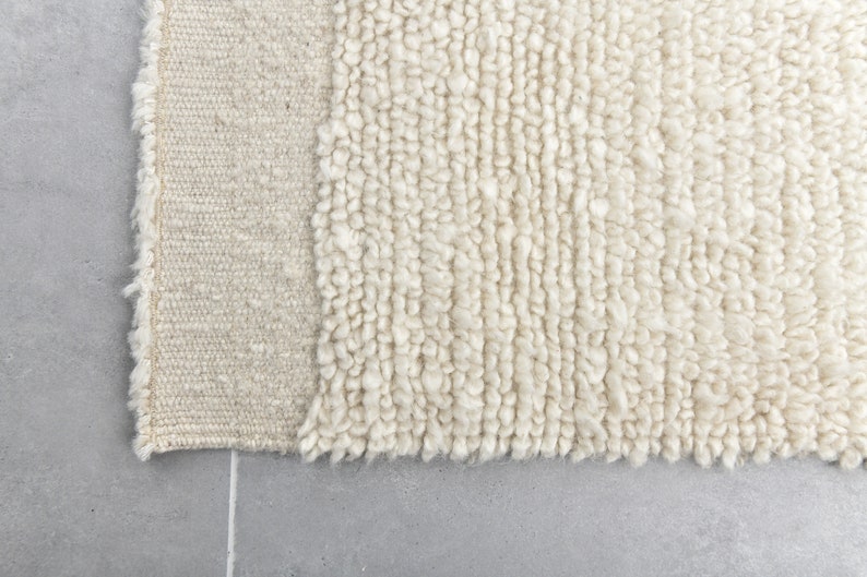 May include: Close-up of a cream-colored rug with a textured, fluffy surface. The rug has a woven edge.
