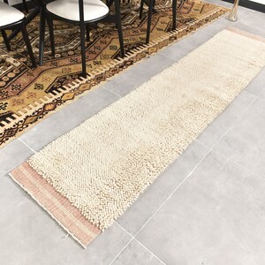 Puede incluir: Una alfombra de pelo blanco y crema con un borde rosa. La alfombra est&aacute; colocada sobre un suelo de baldosas grises.