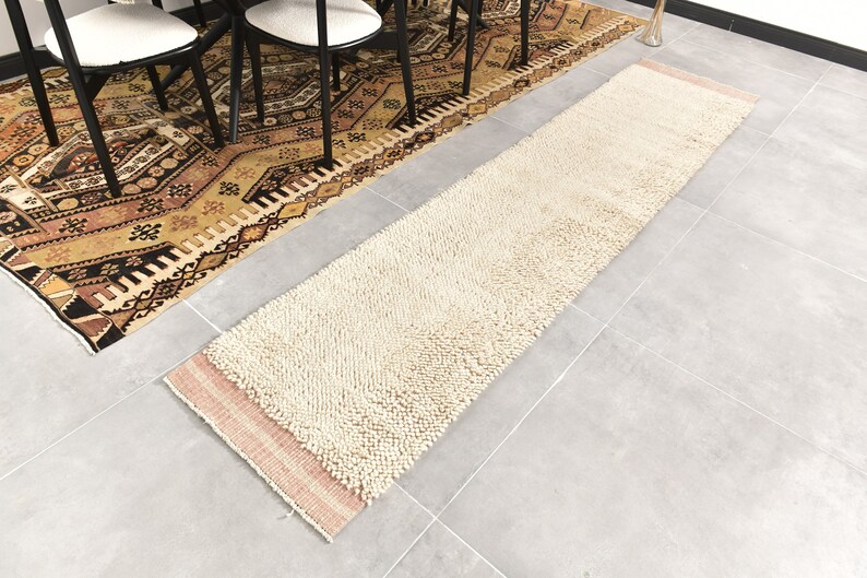Puede incluir: Una alfombra de pelo blanco y crema con un borde rosa. La alfombra est&aacute; colocada sobre un suelo de baldosas grises.