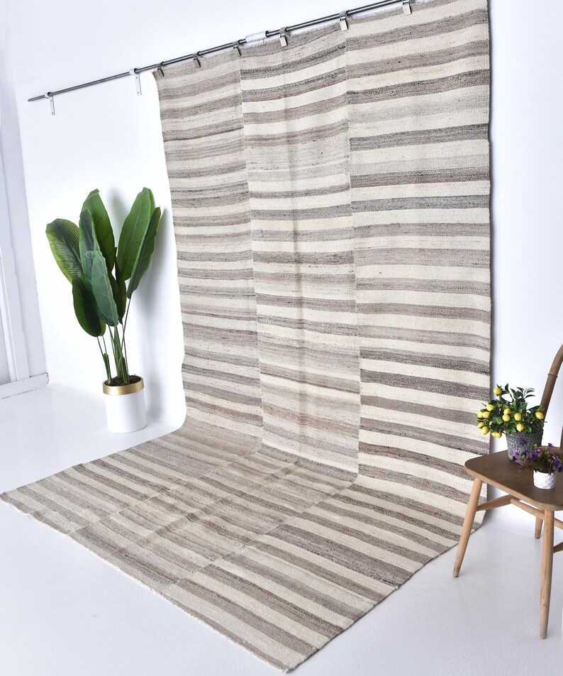 Pu&ograve; includere: Un grande tappeto a strisce in tonalit&agrave; beige e bianco &egrave; appeso a un muro. Il tappeto &egrave; realizzato in fibre naturali e ha un aspetto rustico e tessuto a mano.