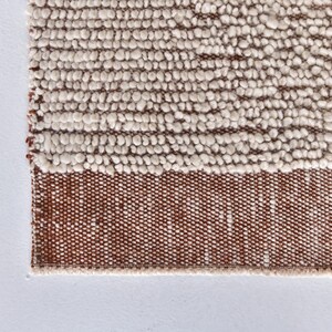 May include: Close-up of a white and brown woven rug with a textured surface. The rug has a fringe border.