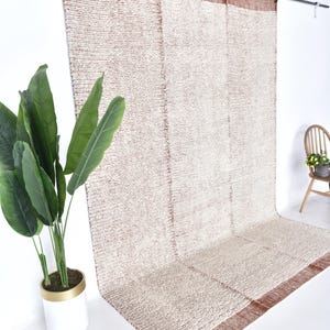 May include: A white and rust-colored woven rug hangs on a wall. The rug has a textured, fluffy surface and a subtle pattern. The rug is hanging on a white wall with a plant in a white pot in the foreground.