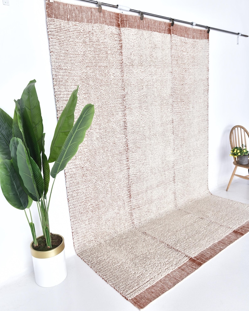 May include: A white and rust-colored woven rug hangs on a wall. The rug has a textured, fluffy surface and a subtle pattern. The rug is hanging on a white wall with a plant in a white pot in the foreground.