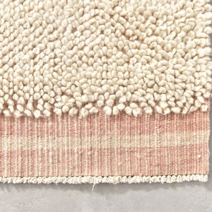 Puede incluir: Una alfombra de color crema con una textura esponjosa y un borde rayado rosa y crema. La alfombra tiene un borde de flecos.