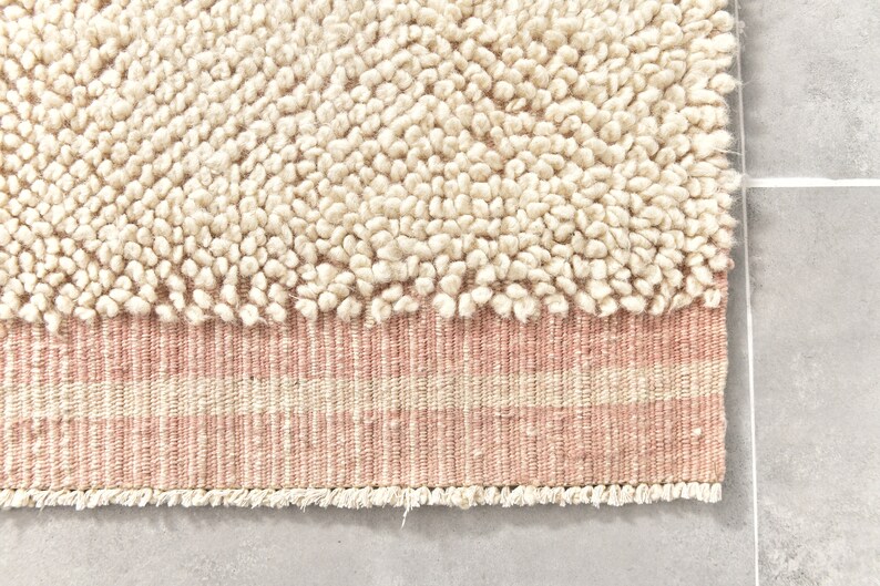 Puede incluir: Una alfombra de color crema con una textura esponjosa y un borde rayado rosa y crema. La alfombra tiene un borde de flecos.